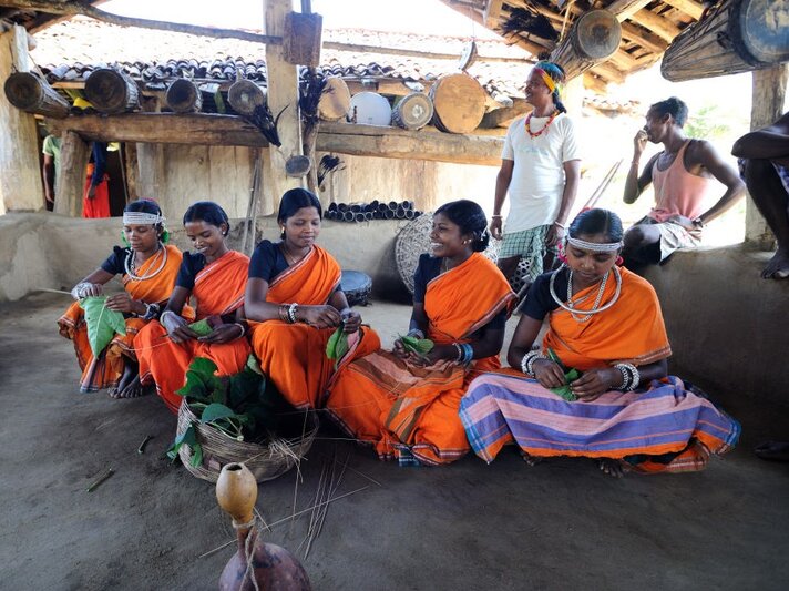 Sechs junge Adivasi-Frauen in orangefarbenen Saris sitzen lächelnd auf dem Boden und falten Blätter in einer offenen Lehmhütte. | © Getty Images / Frédéric Soltan