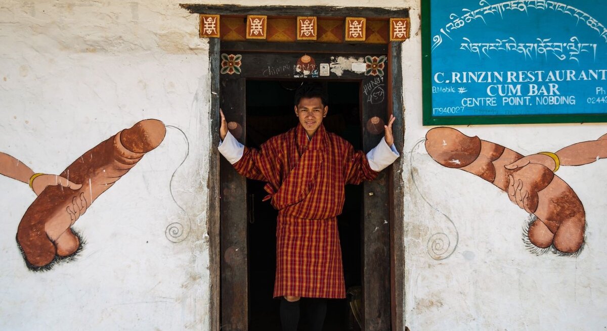 Junger Bhutaner in rot-gelbem Gho steht lächelnd im Türrahmen, flankiert von traditionellen Phallusmalereien an weißer Wand.