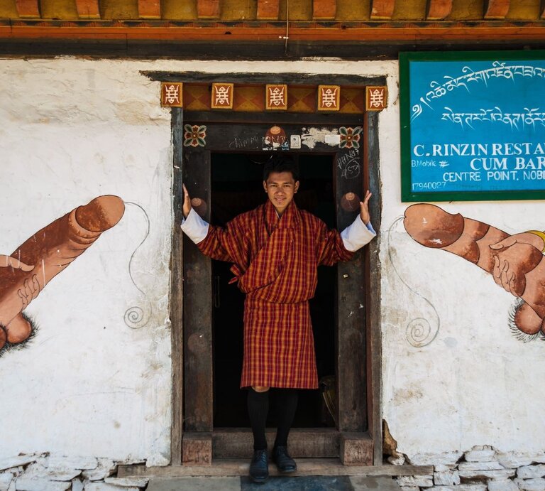Junger Bhutaner in rot-gelbem Gho steht lächelnd im Türrahmen, flankiert von traditionellen Phallusmalereien an weißer Wand. | © Getty Images / David DUCOIN