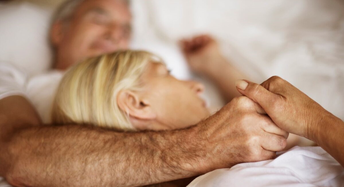 Ein älteres Paar liegt eng umschlungen in einem weißen Bett, hält sich liebevoll an den Händen und wirkt entspannt und geborgen.