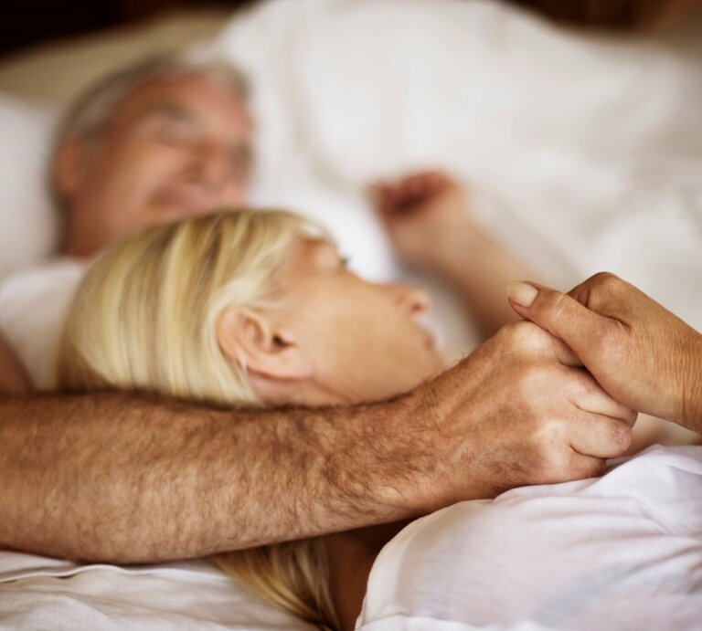 Ein älteres Paar liegt eng umschlungen in einem weißen Bett, hält sich liebevoll an den Händen und wirkt entspannt und geborgen. | © Getty Images / Solskin