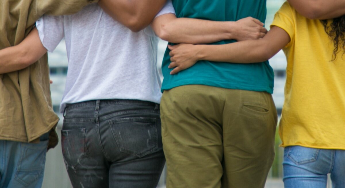Gruppe von vier Personen, die einander umarmen, von hinten betrachtet. Jeder hält eine andere Person liebevoll um die Schultern oder den Rücken.