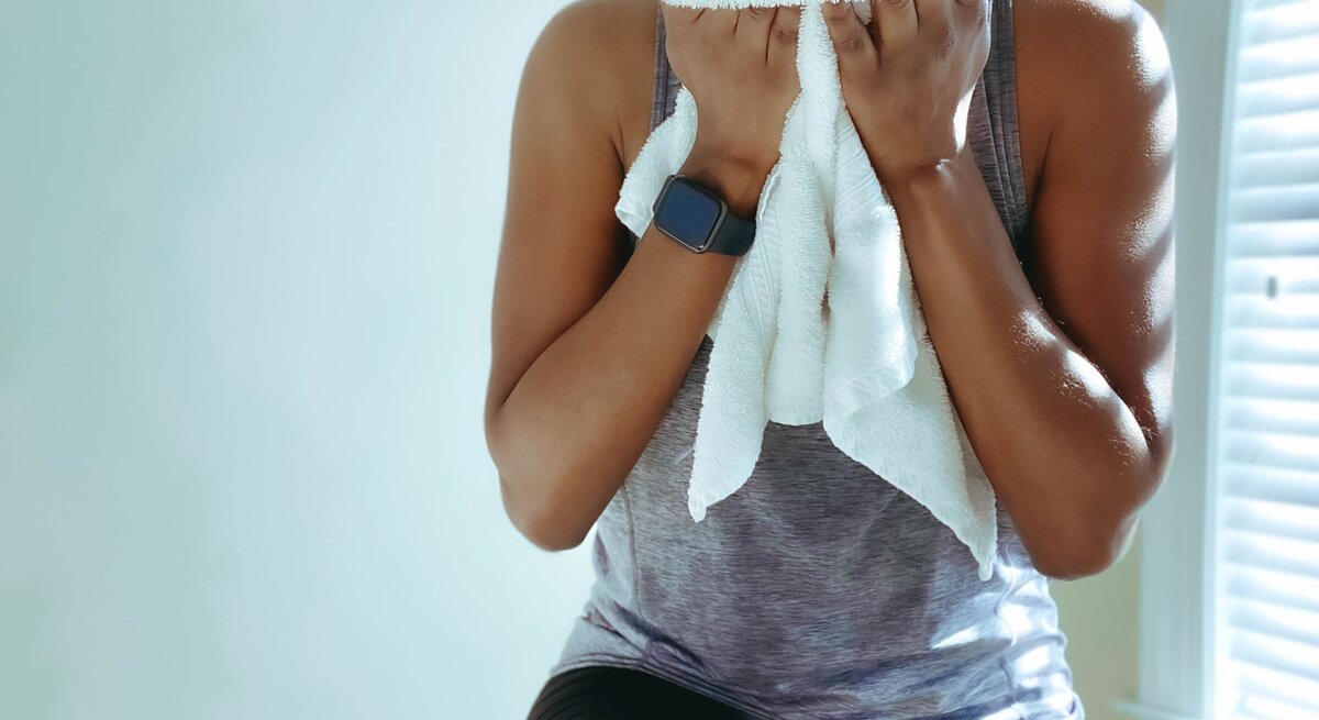 Eine sportliche Frau in grauem Tanktop wischt sich mit einem weißen Handtuch das Gesicht, Sonnenlicht fällt durch ein Fenster.
