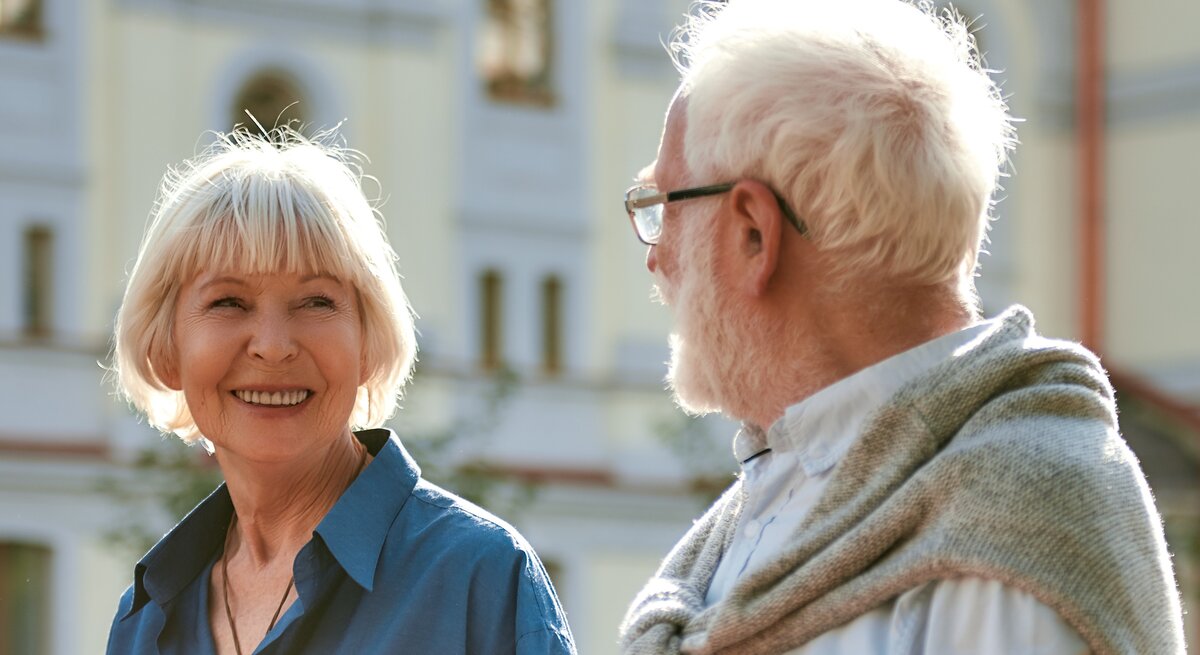 Eine ältere Frau mit weißem Bob lächelt fröhlich einen weißhaarigen Mann mit Brille an, im Hintergrund eine sonnige Gebäudefassade.