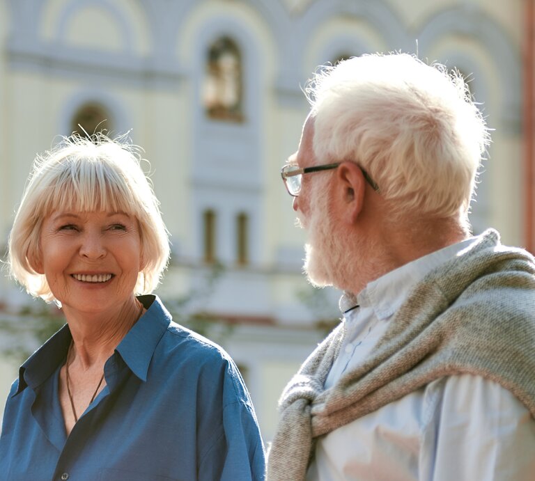 Eine ältere Frau mit weißem Bob lächelt fröhlich einen weißhaarigen Mann mit Brille an, im Hintergrund eine sonnige Gebäudefassade. | © Adobe Stock / Kostiantyn