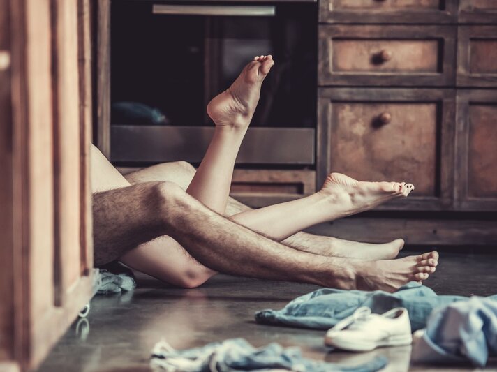 Zwei ineinander verschlungene Beine eines Paares liegen auf einem Küchenboden, umgeben von verstreuten Kleidungsstücken und Schuhen. | © Adobe Stock / VadimGuzhva