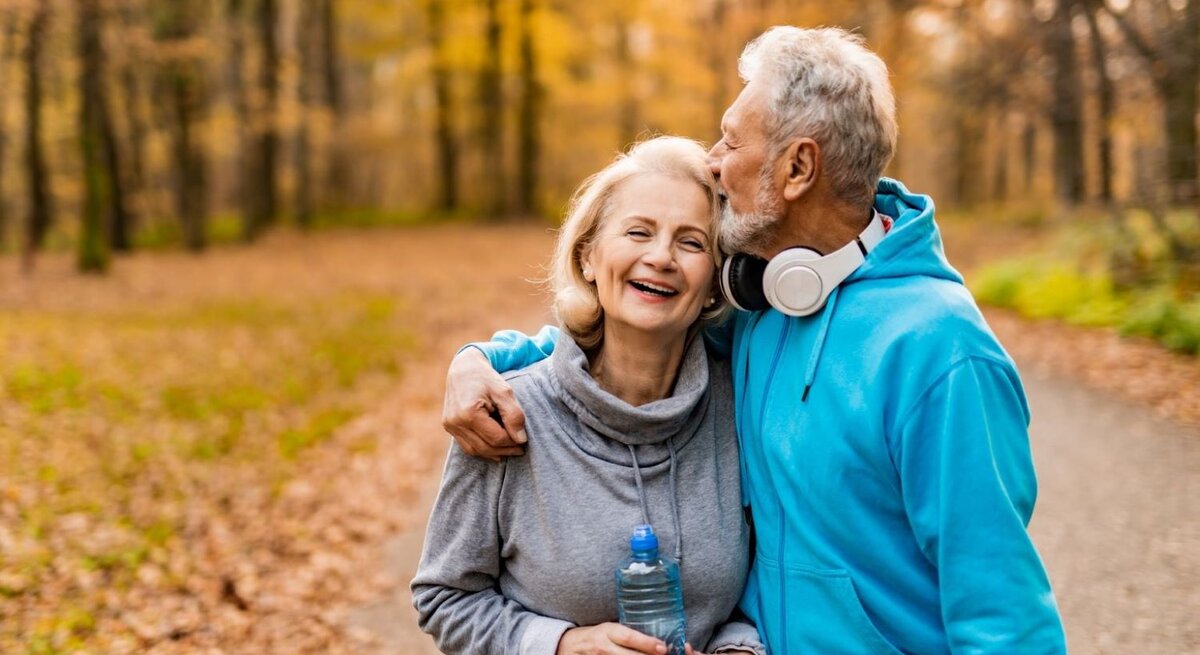 Ein älteres Paar spaziert lachend durch einen herbstlichen Wald, der Mann in blauer Sportkleidung küsst die Frau auf die Stirn.