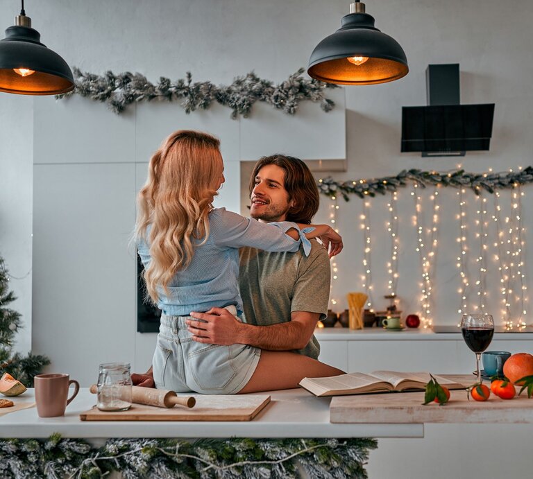 Ein Paar sitzt verliebt in einer modern dekorierten Küche, mit einem geschmückten Weihnachtsbaum und Lichterketten im Hintergrund. | © Getty Images/ Harbucks