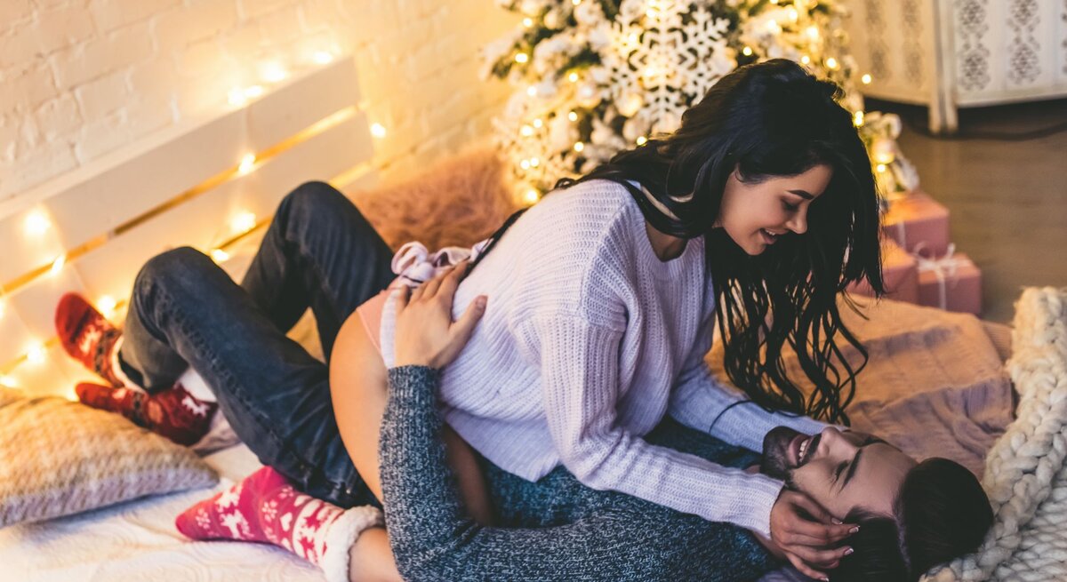 Ein Paar kuschelt verliebt auf einem Bett mit Lichterketten und Weihnachtsbaum im Hintergrund, beide wirken entspannt und fröhlich.