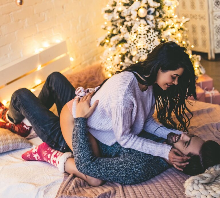 Ein Paar kuschelt verliebt auf einem Bett mit Lichterketten und Weihnachtsbaum im Hintergrund, beide wirken entspannt und fröhlich. | © Getty Images/ Vasyl Dolmatov