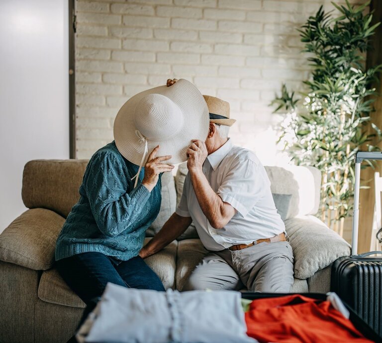 Älteres Paar, das augenscheinlich Spaß hat, während es auf einem Sofa sitzt und sich hinter einem weißen Sommerhut zu küssen scheint. | © iStock.com/supersizer