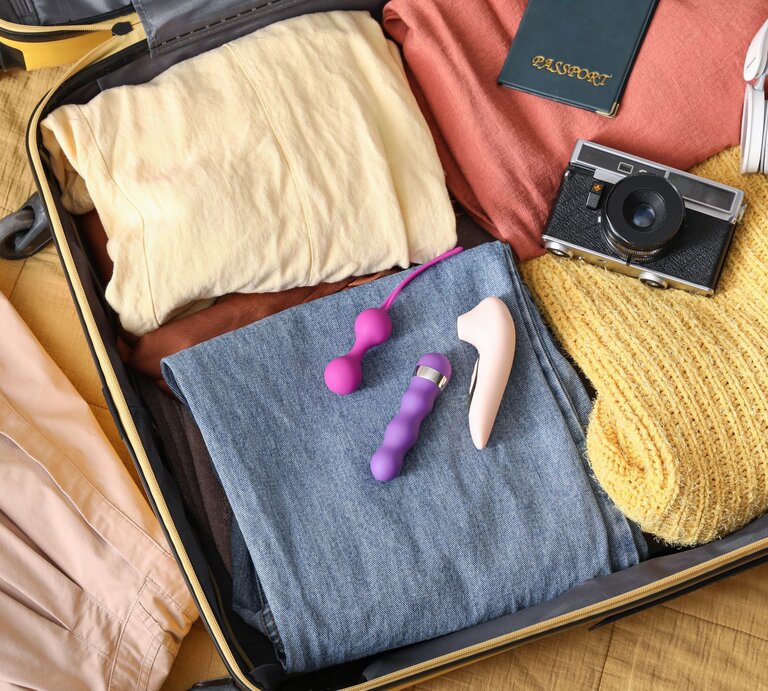 Offener Koffer auf einem Bett mit gefalteten Kleidungsstücken, Kamera, Pass, Kopfhörern und lilafarbenen Sexspielzeugen. | © Adobe Stock/ Pixel-Shot