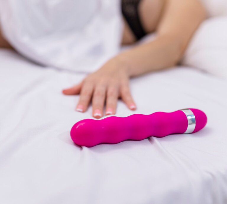 Ein pinkfarbener Vibrator liegt auf einem Bett, eine Frau liegt daneben, von der man nur den Arm sieht, der sich nach dem Sex Toy streckt | © Adobe Stock / руслан малыш