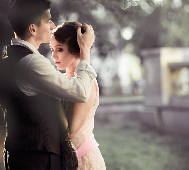 Ein junger Mann in Weste hält sanft den Kopf einer jungen Frau in rosa Kleid, während sie verträumt in einem Park steht. | © Adobe Stock / daryakomarova