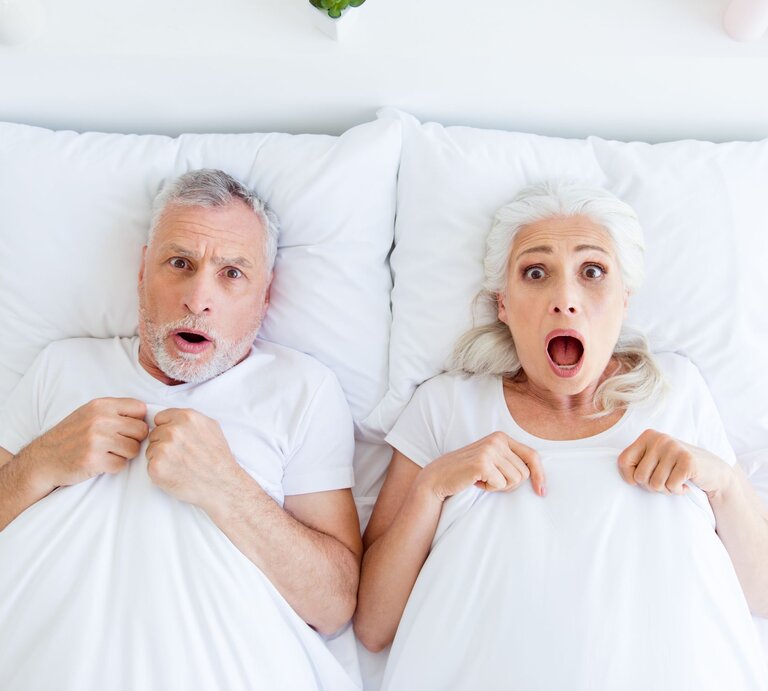 Ein älteres Paar mit weißen Haaren liegt erschrocken im Bett, hält die Decke fest und blickt mit offenen Mündern nach vorn. | © Adobe Stock / deagreez