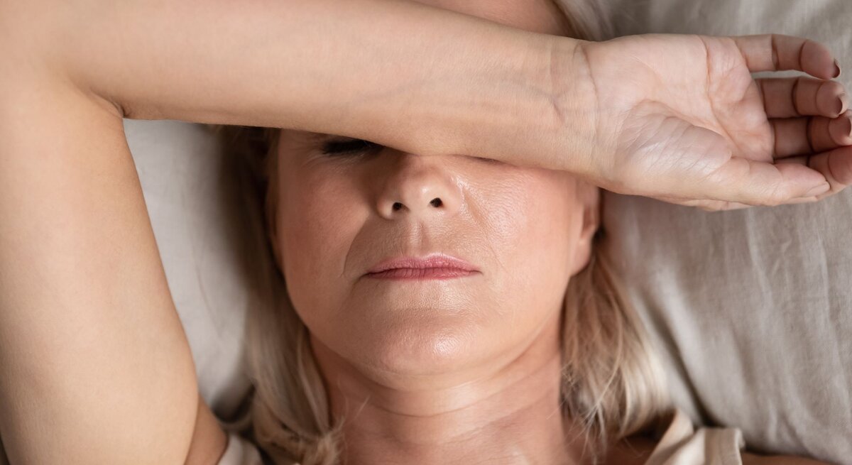 Ältere Frau mit blondem Haar liegt mit ernster Miene im Bett, trägt ein beiges Top und bedeckt ihre Augen mit dem rechten Unterarm.