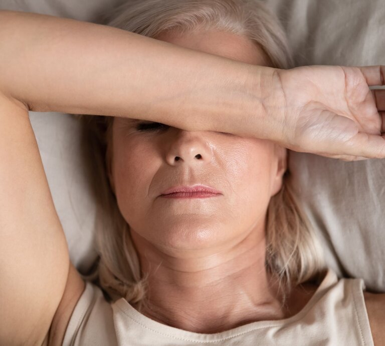 Ältere Frau mit blondem Haar liegt mit ernster Miene im Bett, trägt ein beiges Top und bedeckt ihre Augen mit dem rechten Unterarm. | © Adobe Stock / fizkes