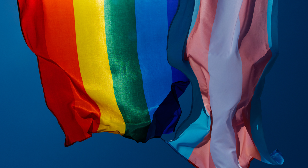 Regenbogen- und Transgender-Flagge wehen nebeneinander in kräftigen Farben vor klarem, tiefblauem Himmel.