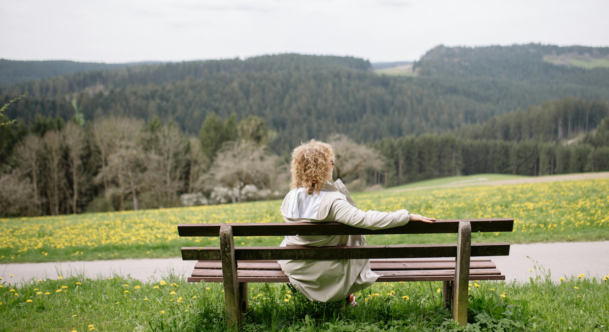 Eine Frau mit lockigem Haar und weißem Mantel sitzt entspannt auf einer Holzbänken, blickt auf eine blühende Frühlingswiese und Waldlandschaft.