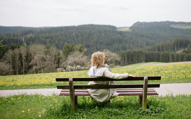 Eine Frau mit lockigem Haar und weißem Mantel sitzt entspannt auf einer Holzbänken, blickt auf eine blühende Frühlingswiese und Waldlandschaft. | © Canva