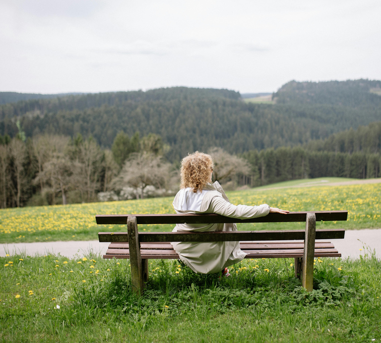 Eine Frau mit lockigem Haar und weißem Mantel sitzt entspannt auf einer Holzbänken, blickt auf eine blühende Frühlingswiese und Waldlandschaft. | © Canva