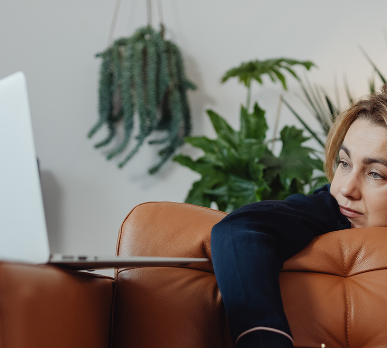 Eine Person lehnt mit dunkelblauem Ärmel entspannt auf einem braunen Ledersofa, im Hintergrund grüne Zimmerpflanzen und ein Laptop. | © Canva