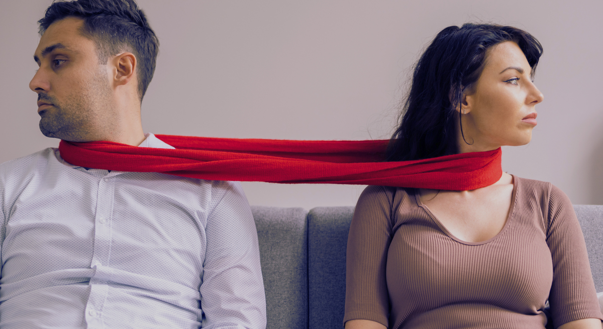 Eine Frau in braunem Shirt und ein Mann in weißem Hemd sitzen auf einem grauen Sofa, verbunden durch einen roten Schal.