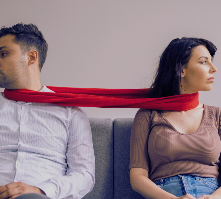 Eine Frau in braunem Shirt und ein Mann in weißem Hemd sitzen auf einem grauen Sofa, verbunden durch einen roten Schal. | © Canva