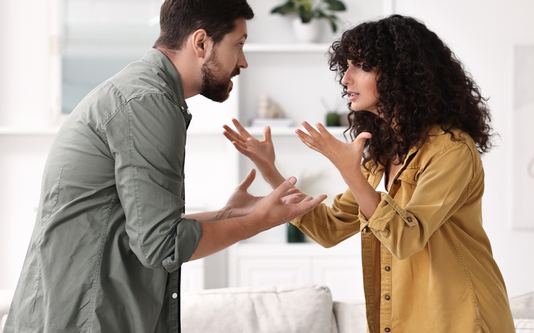 Ein junger Mann mit Bart und eine junge Frau mit Locken streiten gestikulierend im hellen Wohnzimmer mit Pflanze im Hintergrund. | © Canva