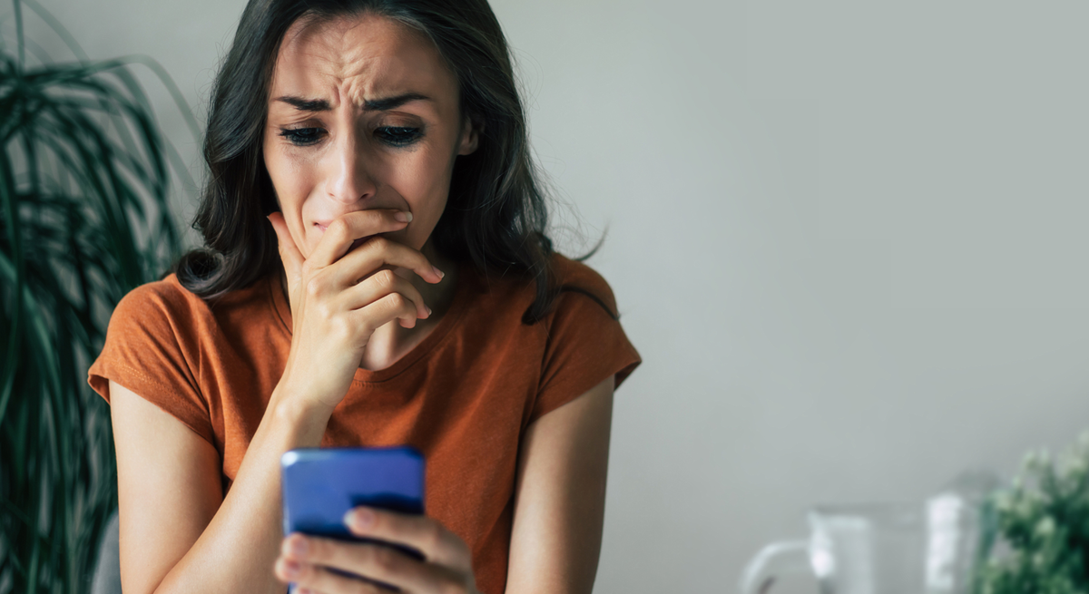 Besorgte Frau in orangefarbenem Shirt liest fassungslos eine Nachricht auf dem Smartphone, im Hintergrund grüne Zimmerpflanze.