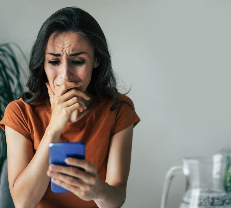 Besorgte Frau in orangefarbenem Shirt liest fassungslos eine Nachricht auf dem Smartphone, im Hintergrund grüne Zimmerpflanze. | © Canva