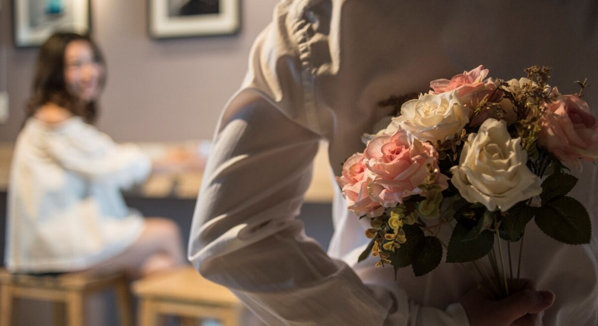 Ein Mann in weißem Hemd versteckt einen Rosenstrauß hinter dem Rücken, während eine junge Frau ihn lächelnd ansieht.