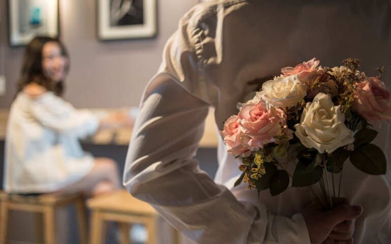 Ein Mann in weißem Hemd versteckt einen Rosenstrauß hinter dem Rücken, während eine junge Frau ihn lächelnd ansieht. | © Getty Images / Nattachart Jerdnapapunt