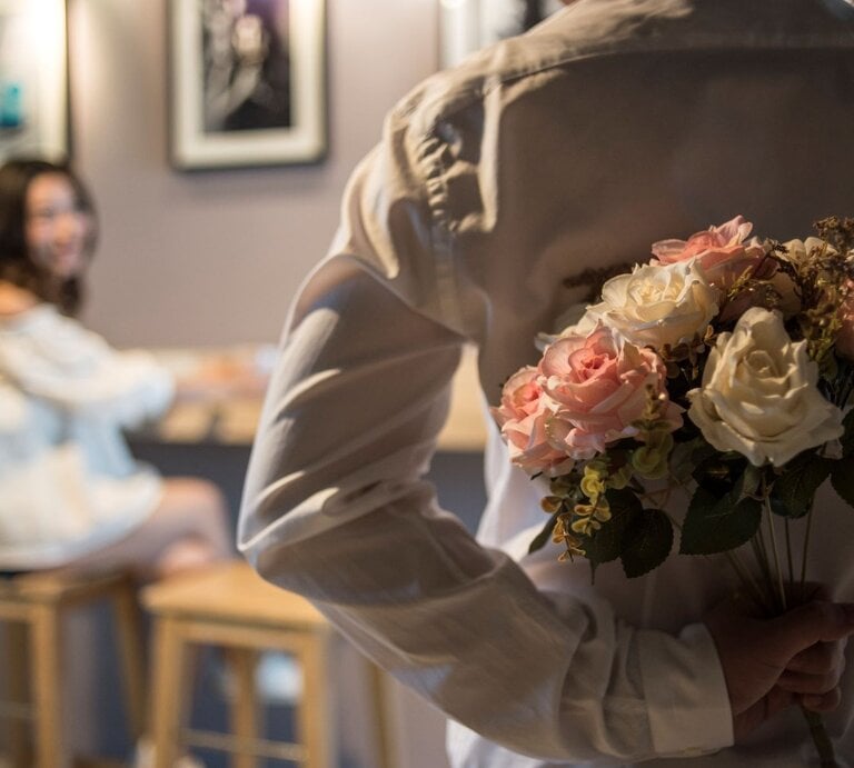 Ein Mann in weißem Hemd versteckt einen Rosenstrauß hinter dem Rücken, während eine junge Frau ihn lächelnd ansieht. | © Getty Images / Nattachart Jerdnapapunt