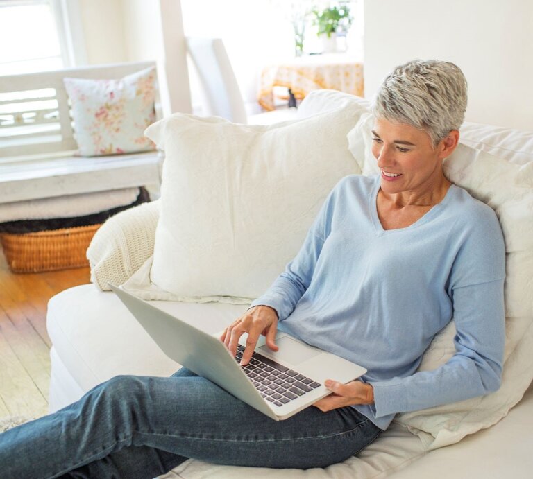 Lächelnde grauhaarige Frau in Jeans und hellblauem Pullover liegt entspannt mit Laptop auf weißem Sofa in heller, gemütlicher Wohnung. | © Getty Images / Jamie Kingham