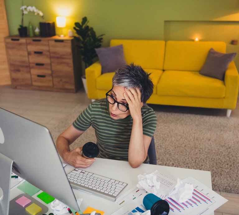Eine gestresste Frau mit kurzer grauer Frisur sitzt mit Kaffeebecher vor Papieren am Schreibtisch in einem bunten Wohnzimmer. | © Adobe Stock / deagreez