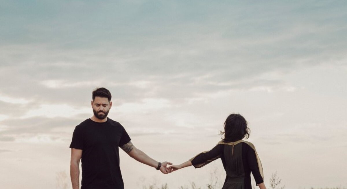 Ein bärtiger Mann in Schwarz hält traurig die Hand einer Frau im Kleid, die sich abwendet, vor wolkigem Himmel auf dem Feld.