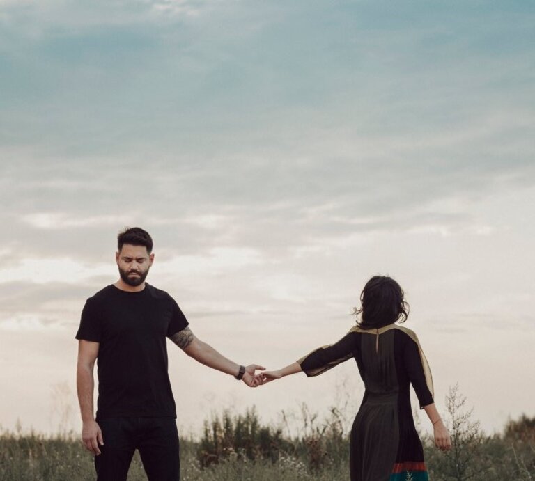 Ein bärtiger Mann in Schwarz hält traurig die Hand einer Frau im Kleid, die sich abwendet, vor wolkigem Himmel auf dem Feld. | © iStock/proud_natalia