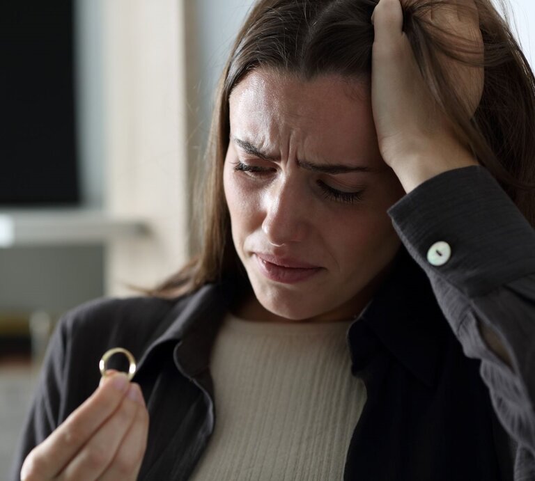 Junge Frau mit langen braunen Haaren blickt traurig auf einen Ring in der Hand, stützt den Kopf verzweifelt mit der anderen. | © Getty Images / AntonioGuillem
