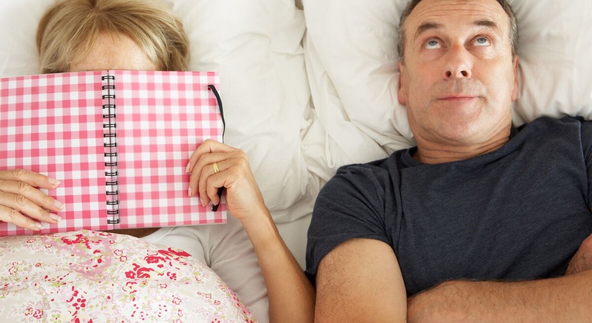 Mittelaltes Paar liegt im Bett, die Frau verbirgt ihr Gesicht hinter einem karierten Notizbuch, der Mann blickt genervt zur Decke.