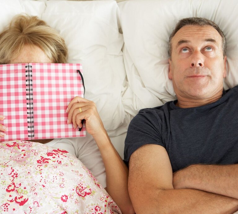 Mittelaltes Paar liegt im Bett, die Frau verbirgt ihr Gesicht hinter einem karierten Notizbuch, der Mann blickt genervt zur Decke. | © Getty images / monkeybusinessimages