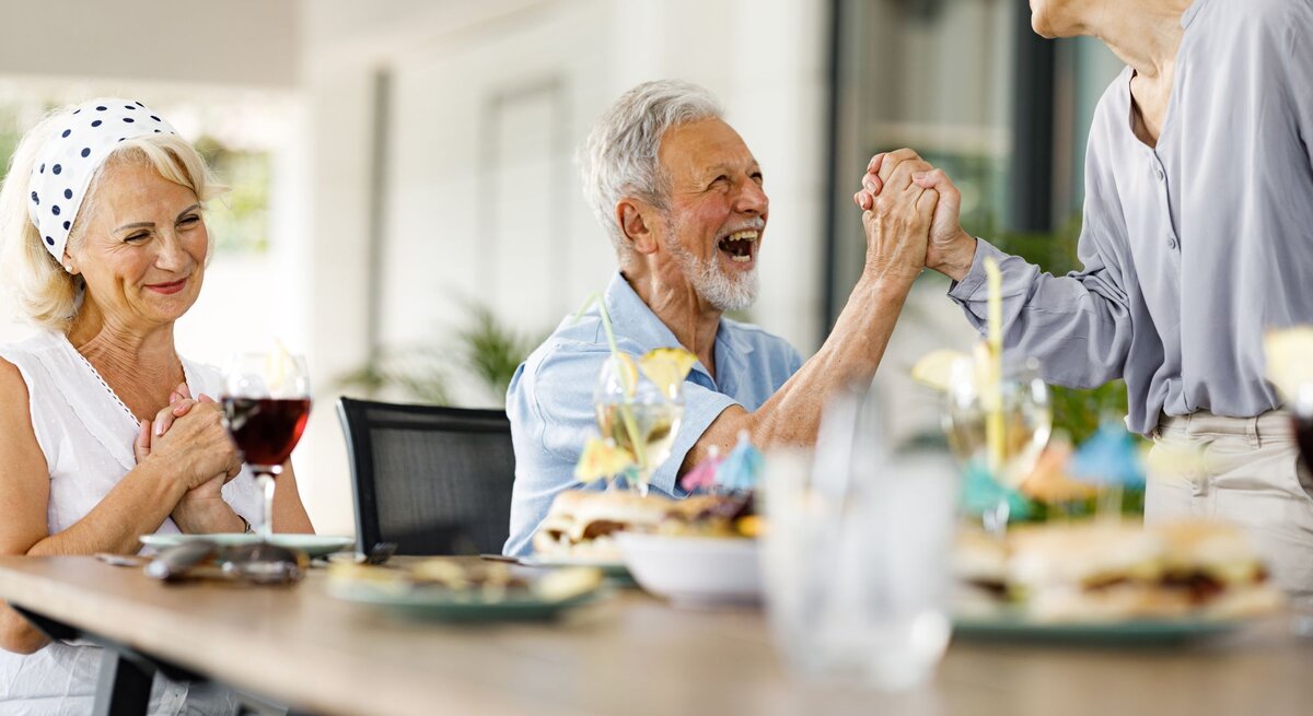 Drei ältere Menschen lachen und feiern an einem gedeckten Tisch im Freien, während zwei sich herzlich die Hände reichen.