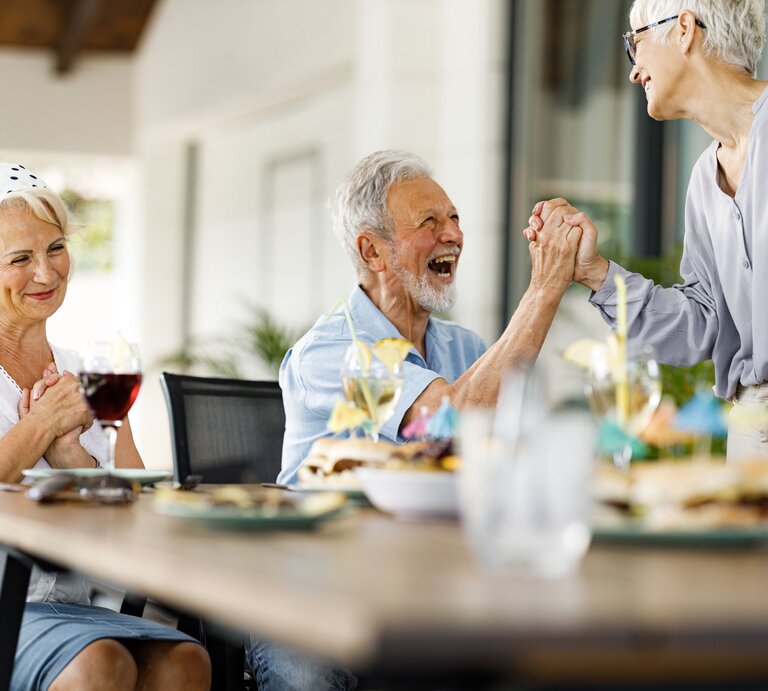 Drei ältere Menschen lachen und feiern an einem gedeckten Tisch im Freien, während zwei sich herzlich die Hände reichen. | © Getty Images / skynesher