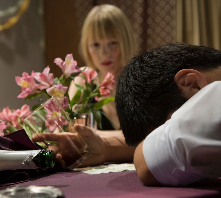 Ein Mann in weißem Hemd liegt mit dem Kopf auf einem Tisch, ein leeres Glas in der Hand, während eine blonde Frau ihn ernst ansieht. | © Getty Images / Hill Street Studios