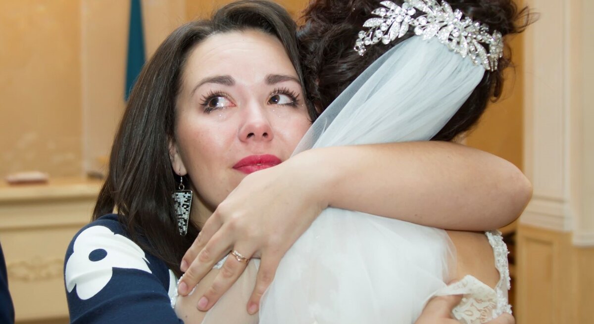 Eine Braut mit funkelndem Haarschmuck und Schleier umarmt eine weinende Frau in einem dunkelblauen Kleid mit weißen Mustern.
