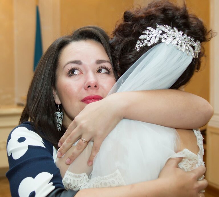 Eine Braut mit funkelndem Haarschmuck und Schleier umarmt eine weinende Frau in einem dunkelblauen Kleid mit weißen Mustern. | © Getty Images / Sviatlana Lazarenka