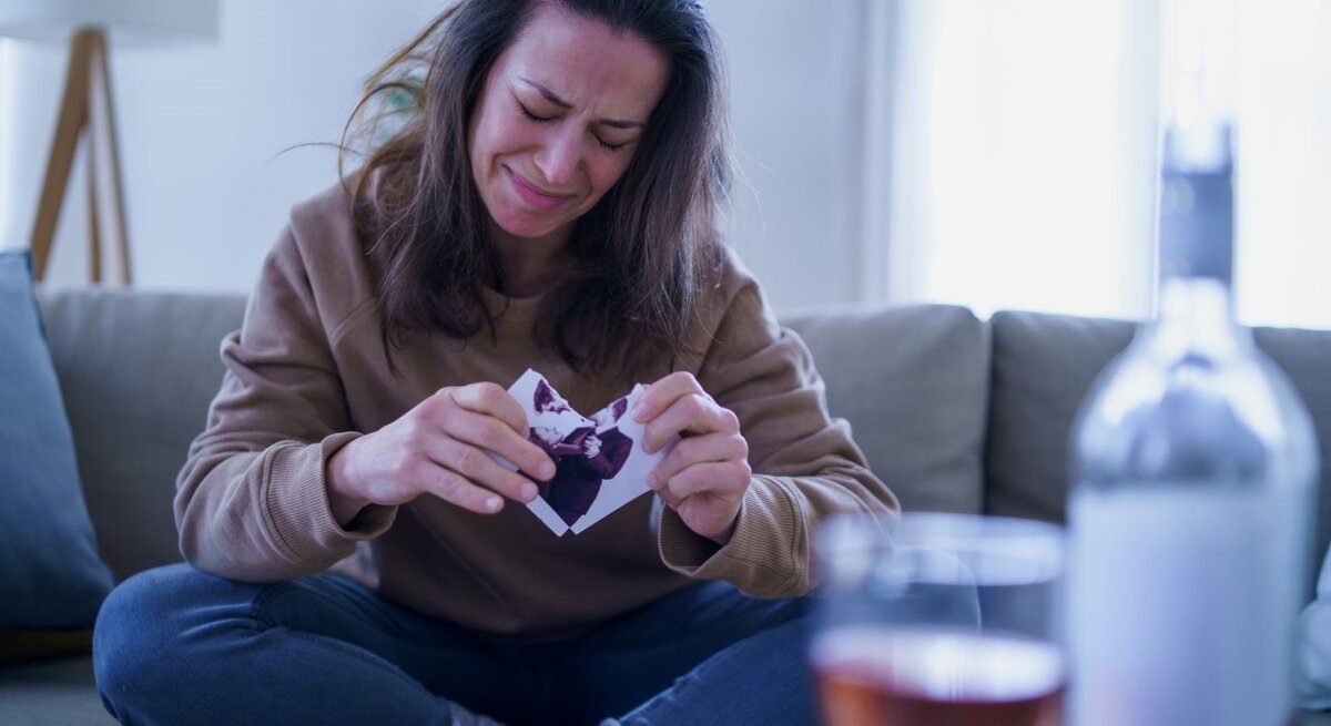 Eine junge Frau mit braunen Haaren sitzt weinend auf dem Sofa und zerreißt ein Foto, vor ihr eine geöffnete Weinflasche und ein Glas.