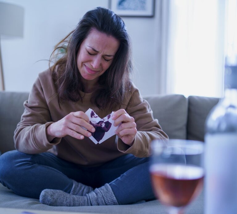 Eine junge Frau mit braunen Haaren sitzt weinend auf dem Sofa und zerreißt ein Foto, vor ihr eine geöffnete Weinflasche und ein Glas. | © Getty Images / Halfpoint Images