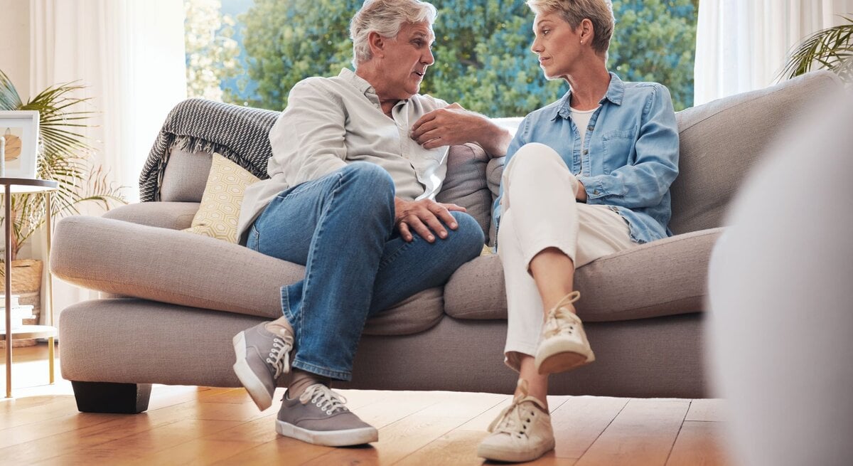 Ein älteres Paar sitzt auf einem beigen Sofa im sonnendurchfluteten Wohnzimmer und führt ein ernstes Gespräch.