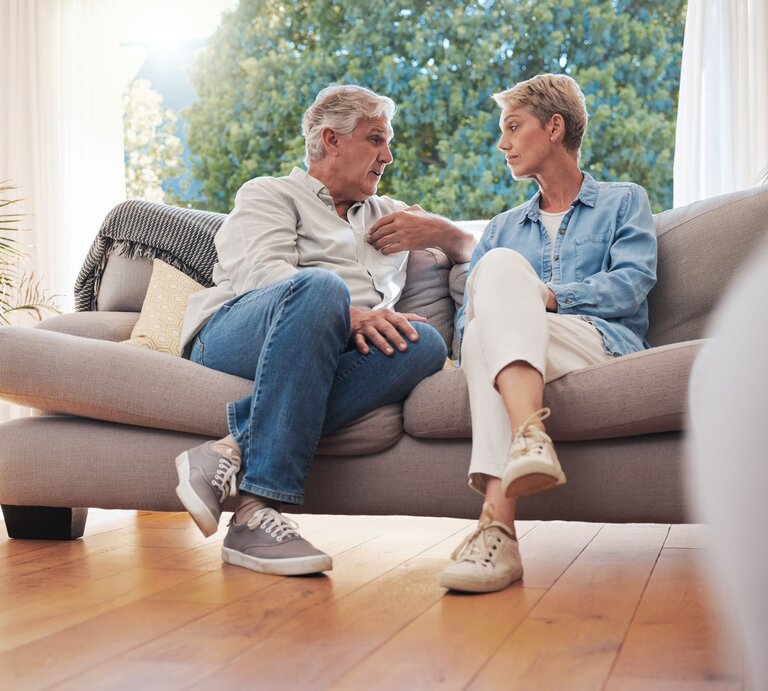 Ein älteres Paar sitzt auf einem beigen Sofa im sonnendurchfluteten Wohnzimmer und führt ein ernstes Gespräch. | © Adobe Stock / Kirsten Davis/peopleimages.com